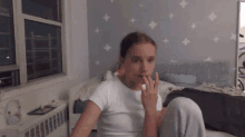 a girl in a white shirt is sitting on a bed with her hand in her mouth