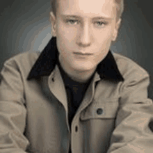 a young man wearing a tan jacket and a black shirt is sitting down .