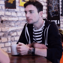 a man in a black and white striped shirt is sitting at a table holding a cup of coffee .