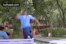 a man in a blue shirt is standing on a bench in a park with his hands in the air .