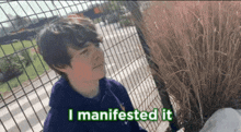 a boy is standing in front of a fence with the words " i manifested it " on the bottom