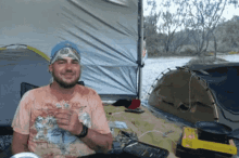 a man with a bandana on his head is sitting in front of a tent holding a glass of water