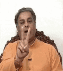 a man in an orange shirt is sitting in a chair and making a funny face with his finger in his mouth .