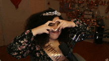 a woman wearing a birthday sash and tiara making a heart shape with her hands