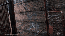 a man in a black shirt is standing in front of a brick wall with a nbc logo on the bottom right