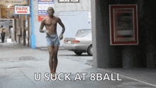 a man without a shirt is running down a street with the words `` u suck at 8ball '' written on the sidewalk .