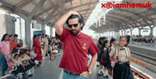 a man wearing sunglasses and a maroon shirt is standing in front of a crowd of people at a train station