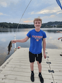 a young man wearing a carmel rams shirt holds a fish on a dock