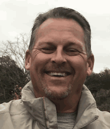 a man with a beard is smiling and wearing a tan jacket