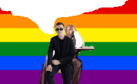 a man in sunglasses sits next to another man in front of a rainbow background