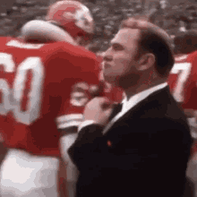 a man in a suit and tie is standing in front of a group of football players in red uniforms .