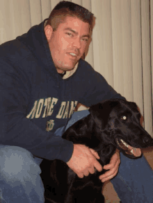 a man wearing a hoodie that says notre day holds a black dog