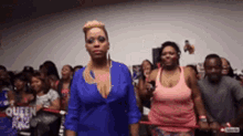 a woman in a blue shirt stands in front of a crowd with the word queen on the wall behind her