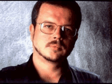 a man with a beard and glasses is standing in front of a wall .