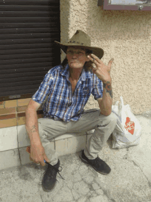 a man wearing a cowboy hat sits on the sidewalk next to a bag that says ' cards price '