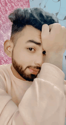 a man with a beard wearing a pink sweater holds his hand to his forehead