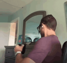 a man in a purple shirt is sitting in front of a mirror holding a remote control .