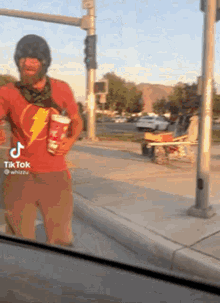 a man in a red shirt with a lightning bolt on it is standing on a sidewalk holding a cup .