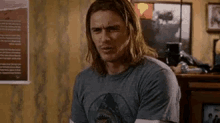 a man with long hair is sitting in a living room wearing a gray t-shirt .