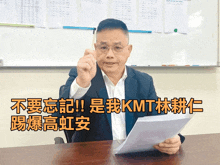 a man in a suit is sitting at a desk holding a piece of paper in front of a whiteboard that says kmt