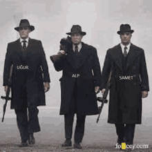 three men in suits and hats are walking down a dirt road with guns .