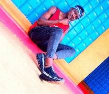 a man wearing a red tank top and black jeans sits on the floor in front of a wall of blue lockers