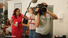 a woman in a red dress is being filmed by a man with a camera in front of a sign that says aloha