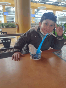 a young boy sits at a table with a cup of ice cream and a blue spoon in it