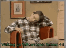 a man in a plaid shirt is sitting in a red chair with the words " waiting on yellowstone season 4 " above him