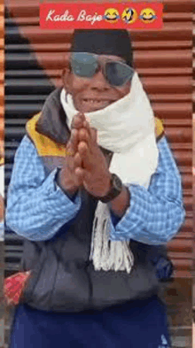 a man wearing sunglasses , a scarf and a hat is standing in front of a wall .