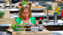 a little girl in a green apron sits at a cutting board with a bowl of food and a bottle of sauce