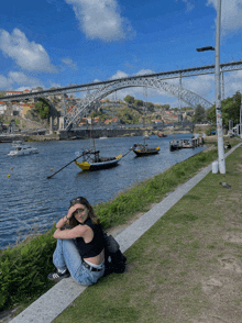 a woman sits on the edge of a river with boats in the water and a bridge in the background