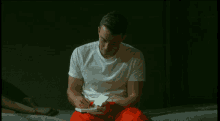 a man in a white t-shirt and red pants is sitting on a bed writing on a piece of paper