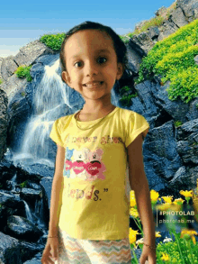 a little girl wearing a yellow shirt that says " never give up " stands in front of a waterfall