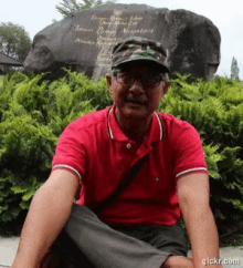 a man wearing a red shirt and a hat sits in front of a large rock that says " taman bunga miniature "