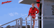 a woman in a red jacket is standing in front of a lifeguard tower with the word baywatch on it