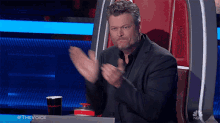 a man in a suit applauds while sitting at a table with a cup of coke in front of him