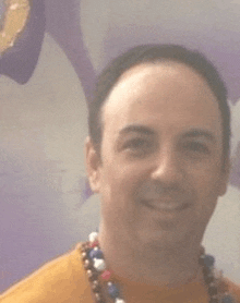 a man wearing a yellow shirt and a necklace of beads is smiling in front of a purple wall .