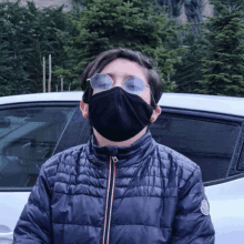 a man wearing a black face mask and sunglasses stands in front of a white car