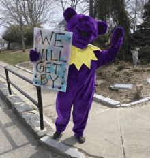 a person in a purple bear costume holds a sign that says we will get by