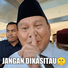 a man wearing a black hat holds his finger to his mouth and says " jangan dikasihu "