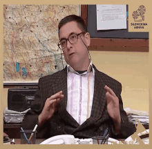 a man wearing glasses and a suit is sitting at a desk in front of a sign that says the best of