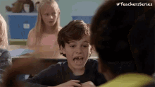 a young boy is sitting at a desk in a classroom with his mouth open and making a funny face .