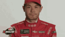 a man wearing a red racing suit and a red hat is standing in front of a nascar sign .