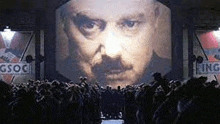 a group of people are standing in front of a large screen displaying a man 's face .