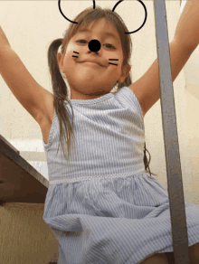 a little girl wearing a blue and white striped dress has a cat face painted on her face