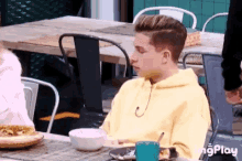 a young man in a yellow hoodie is sitting at a table with a bowl of food and a plate of food .