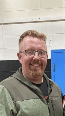 a man with glasses and a beard is smiling in front of a brick wall .