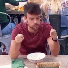 a man in a red shirt is sitting at a table with a bowl of food and a fork .