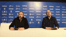 two men sit at a table in front of a wall with betting logos